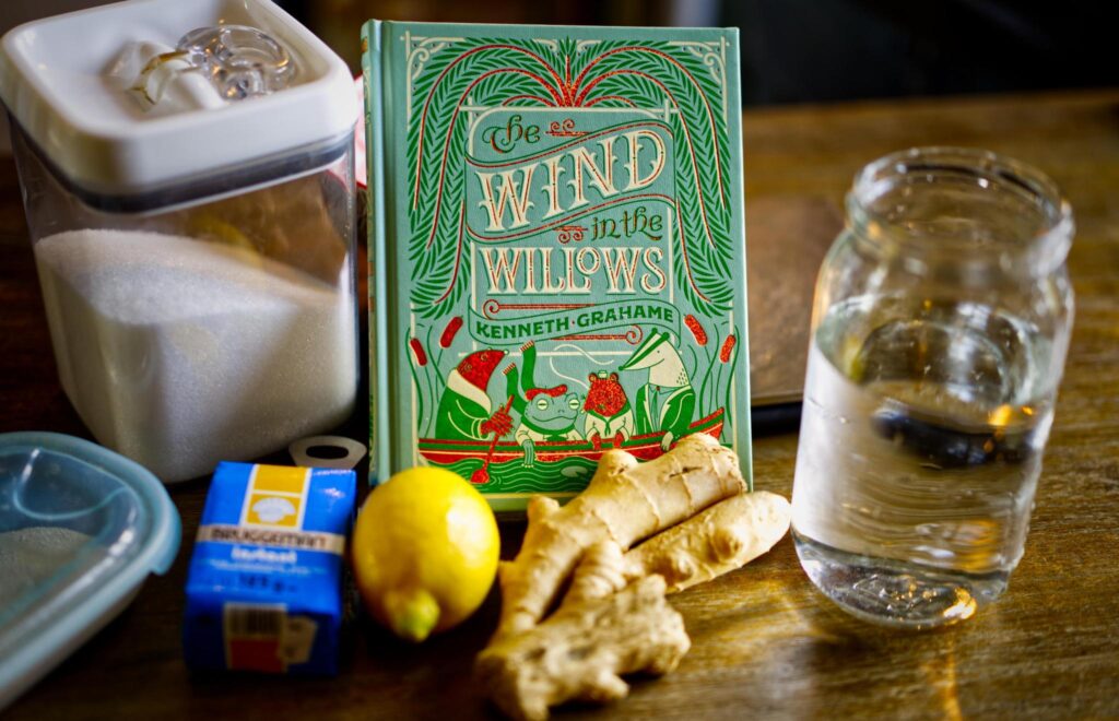 A table with a jar of water, a lemon, a hand of ginger, a contain of yeast, a container of sugar, and a book.