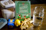 A table with a jar of water, a lemon, a hand of ginger, a contain of yeast, a container of sugar, and a book.