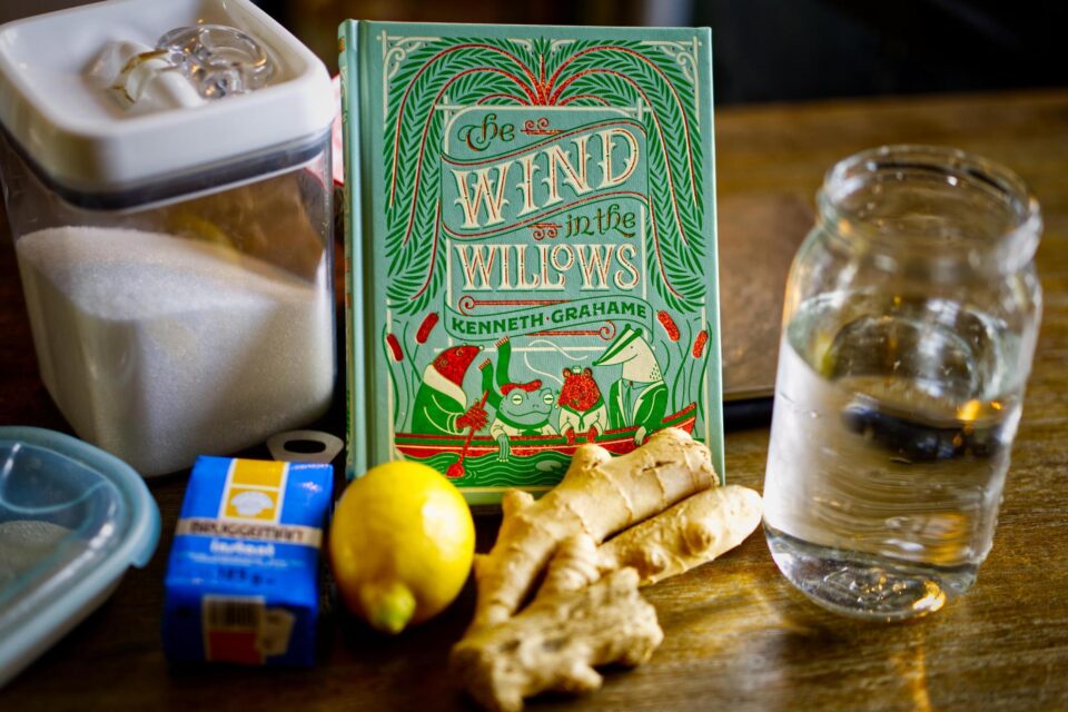 A table with a jar of water, a lemon, a hand of ginger, a contain of yeast, a container of sugar, and a book.