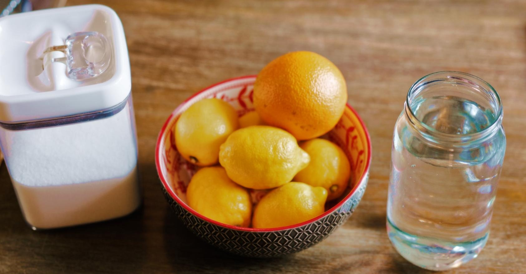 A table with a container of sugar, a bowl with lemons and 1 orange, and a jar of water